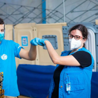 A woman and a man wearing face masks and disposable gloves fist-bump