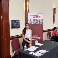 A woman stands and smiles at the camera. In the background there is a roll-up banner for SEMCA, she has in front of her a table with forms.