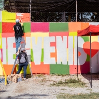 Three people are painting a mural that reads "Bienvenidos"