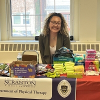 A woman sits at a booth with a Scranton logo. There are menstrual products on the table.