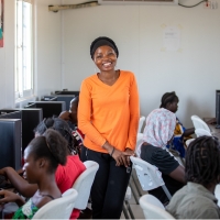A woman smiling and looking at the camera, standing in the middle of a classroom of children using computers