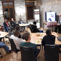 A photo from the event, showing speakers standing while participants sit around small tables in front