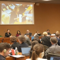Photo from the event: a panel of five people sit facing the audience, while they listen to someone in the audience speaking. The person speaking is shown on a screen behind the panel.