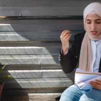 A young woman sitting on a bench, reading from a notebook she is holding in one hand, balanced on her lap. The other hand is raised in expression. Next to her on the bench is a pot plant, and a shaft of sunlight is lights up the wooden panelling of the wall behind her.