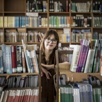 Olha standing in a library, leaning between two bookcases, looking at the camera