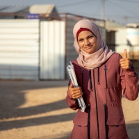 A woman standing outside, with buildings behind her, smiling and giving a thumbs up to the camera. She is holding textbooks and notebooks under one arm