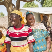 Two women standing together under a tree laughing. There are other people and buildings in the background