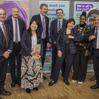 A group photo of nine people smiling and laughing - two of them are putting their elbows together. They are standing in front of banners advertising events at the Global Refugee Forum