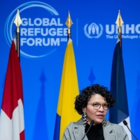 A woman speaking at a podium with eight different flags behind her and the Global Refugee Forum logo on the wall behind the flags