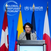 A woman speaking at a podium with eight different flags behind her and the Global Refugee Forum logo on the wall behind the flags