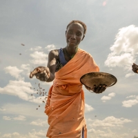 A woman scattering seeds from a metal bowl - the camera is on the ground facing upwards as the seed falls down - the woman is smiling and the sky behind her is bright with a few white fluffy clouds