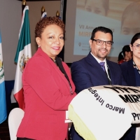 Three people standing together with a folded piece of cloth showing the MIRPS logo with "Presidencia" written on it - behind them are some of the flags of the MIRPS States