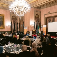 A meeting in what looks like an event room of a grand hotel - there are six round tables around which people are sitting, a stage with a Podium where someone is speaking, and a chandelier coming from the ceiling.