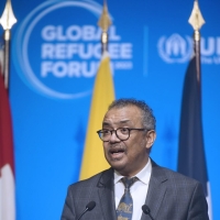 A man standing at a lectern speaking, there are flags behind him with the Global Refugee Forum logo on the wall behind