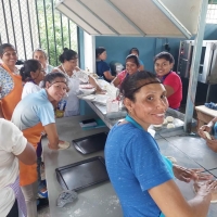 A group of twelve women standing and sitting around a long work table kneading dough.  A couple of them are looking towards the camera, smiling