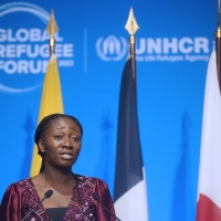 A woman standing at a podium speaking into microphones. Behind her are five flags, and behind the flags a blue background with the Global Refugee Forum logo on it