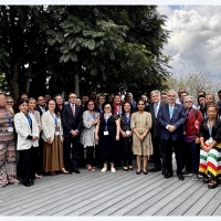 A group photo of around 50 people standing on some wooden decking. There is a tree directly behind those to the left, and behind those on the right is a cloudy, but bright sky.