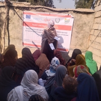 A group of women sitting with one standing at the front handing out leaflets. Behind the one at the front is a banner saying "Health and Hygiene" - they are outside with sand-coloured walls around them