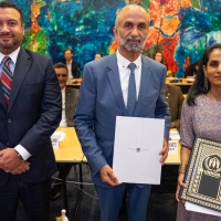 Three people standing next to one another, with a bright abstract painting on the wall behind them. The man in the centre is holding a document with a coat of arms on it. The woman on the right is holding a black and gold plaque with UNHCR's logo on it