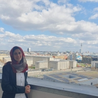 Safia standing on the balcony of German Federal Ministry for Economic Cooperation and Development, behind her is the Berlin skyline, with the TV Tower (Fernsehturm) and Berlin Cathedral (Berliner Dom) in the distance and the Welt hot air balloon in the foreground. The sky is blue with white/light grey clouds.