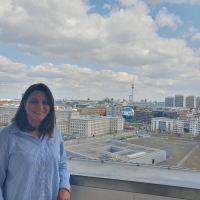 Taban standing on the balcony of German Federal Ministry for Economic Cooperation and Development, behind her is the Berlin skyline, with the TV Tower (Fernsehturm) and Berlin Cathedral (Berliner Dom) in the distance and the Welt hot air balloon in the foreground. The sky is blue with white/light grey clouds.