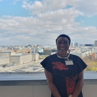 Maryanne standing on the balcony of German Federal Ministry for Economic Cooperation and Development, behind her is the Berlin skyline, with the TV Tower (Fernsehturm) and Berlin Cathedral (Berliner Dom) in the distance and the Welt hot air balloon in the foreground. The sky is blue with white/light grey clouds.