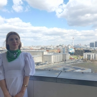 Vlada standing on the balcony of German Federal Ministry for Economic Cooperation and Development, behind her is the Berlin skyline, with the TV Tower (Fernsehturm) and Berlin Cathedral (Berliner Dom) in the distance and the Welt hot air balloon in the foreground. The sky is blue with white/light grey clouds.