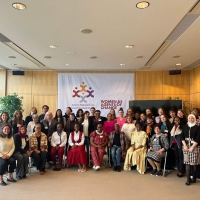 A group photo of participants at the Action Network on Forced Displacement Annual Meeting - around 40 people, those at the front sitting on chairs with others standing behind them. Behind them on a wall is a large banner with the Action Network's logo and the words "Women as Agents of Change"