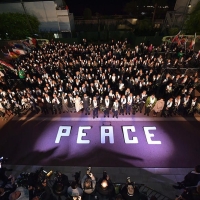An aerial view of a large group of people standing outside, with a purple carpet and word peace written on the ground in front of them. There are flags near the people, and on the other side of the carpet are photographers and videographers
