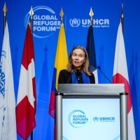 A woman standing at a podium with the Global Refugee Forum logo on it. Behind her are flags on poles, and behind those a blue backdrop with the Global Refugee Forum logo on it.
