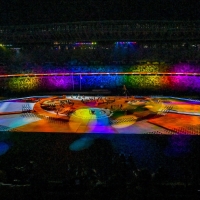 Inside the stadium, it is dark, except for the centre which is lit up with a lot of different coloured bright lights. In the middle, there are people on the stage, and the stands are filled with crowds