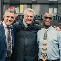 Three men standing together outside on what looks like a street in the centre of a town. The man in the middle has his arms around the shoulders of the other two. They are all smiling
