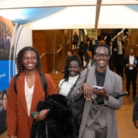 Four people walking through the entry tunnel - an indoor structure within the conference centre with images and the word "Welcome" in different languages on the wall and a tented roof. The four people are smiling at the camera. Behind them more people are descending a staircase coming towards the walk way