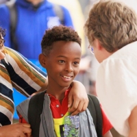 A boy being greeted by a woman, who is bending down to speak to him. He is holding a bottle of water upside down, smiling, while his mother has her arm around his shoulders and is looking at him