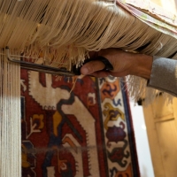 A rug being woven - it is hanging down, and a hand is holding scissors trimming the threads