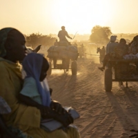 The sun lights up the dust behind groups of Sudanese refugees walking and on carts behind horses