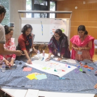 A group of six people standing round a table putting colourful pieces of paper (perhaps some origami?) on a large sheet of paper