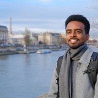 A man standing next to a river. He is smiling at the camera. On the other side of the river are some grand buildings, and in the distance is a boat.