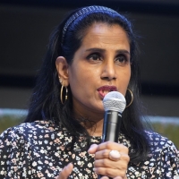 Ruven speaking into a microphone. A close up image only showing her upper body and torso, she is wearing a floral shirt, and holding out one hand to gesture while speaking.