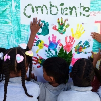 Children painting a picture with their hands. The photo is taken from behind the 5-6 children, and they have made handprints in different colours on a piece of paper, with the word 'ambiente' painted in what looks like fingerprints.