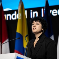A woman standing at a lectern speaking, there are flags and a large screen behind her