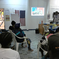 People seating on chairs in a room, listening to someone giving a presentation.