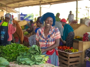 A woman at a vegetable stand in a market smiles at the camera