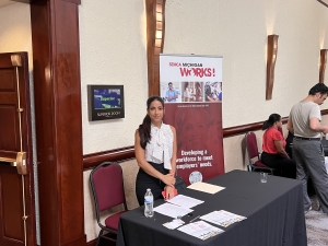 A woman stands and smiles at the camera. In the background there is a roll-up banner for SEMCA, she has in front of her a table with forms.