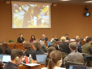 Photo from the event: a panel of five people sit facing the audience, while they listen to someone in the audience speaking. The person speaking is shown on a screen behind the panel.