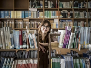 Olha standing in a library, leaning between two bookcases, looking at the camera