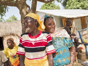 Two women standing together under a tree laughing. There are other people and buildings in the background