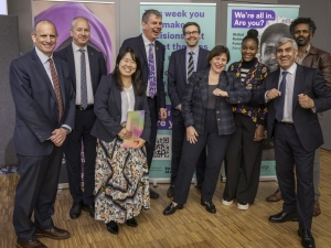 A group photo of nine people smiling and laughing - two of them are putting their elbows together. They are standing in front of banners advertising events at the Global Refugee Forum