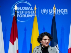 A woman speaking at a podium with eight different flags behind her and the Global Refugee Forum logo on the wall behind the flags