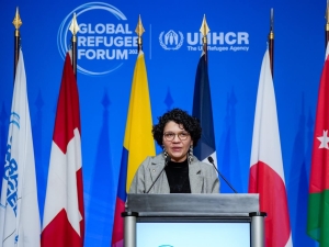 A woman speaking at a podium with eight different flags behind her and the Global Refugee Forum logo on the wall behind the flags